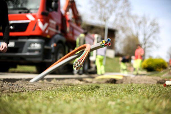 Eine Baustelle bei der Glasfaserkabel aus der Erde schauen. Im Hintergrund Arbeiter sowie ein LKW.